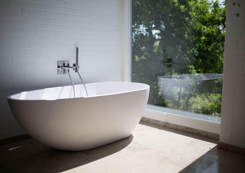 A clean, white bathtub is pictured in front of a white brick wall and large window.