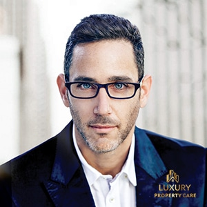 light-skinned man with dark hair, a dark blue suit, blue eyes, slight stubble, short, dark hair, and black rectangular glasses